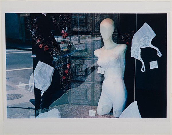 Color photograph looking into the window display of a store featuring women's undergarments.  Partial mannequin featured prominently to the right.  Reflections of the street seen on the left.