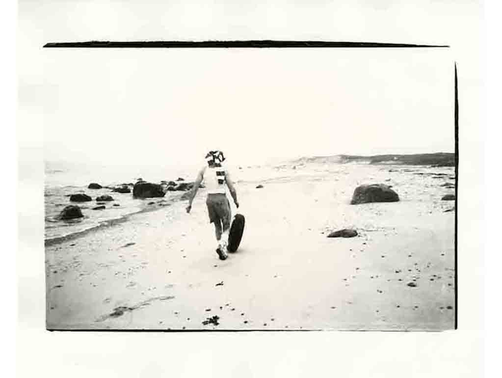 Black and white photograph showing the back of a man as he walks along a beach following a tire.