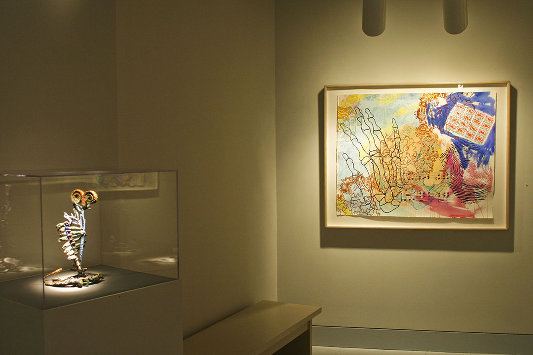 Print and sculpture by Nancy Graves in a gallery.  Sculpture to left in a case. Print shows a skeletal hand, fingerprint, musical notes over other forms