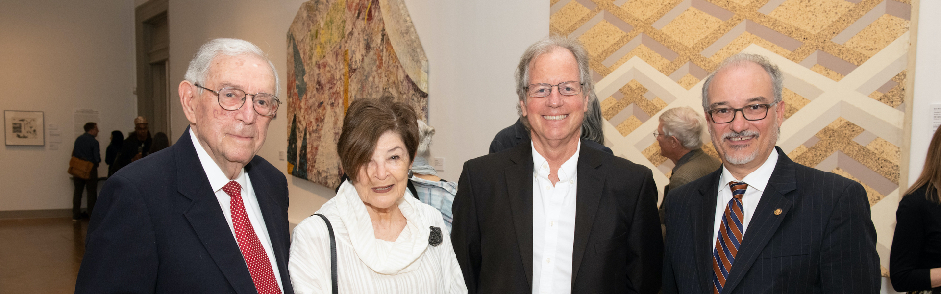 4 people smiling in front of paintings during an event, other people can be seen behind them