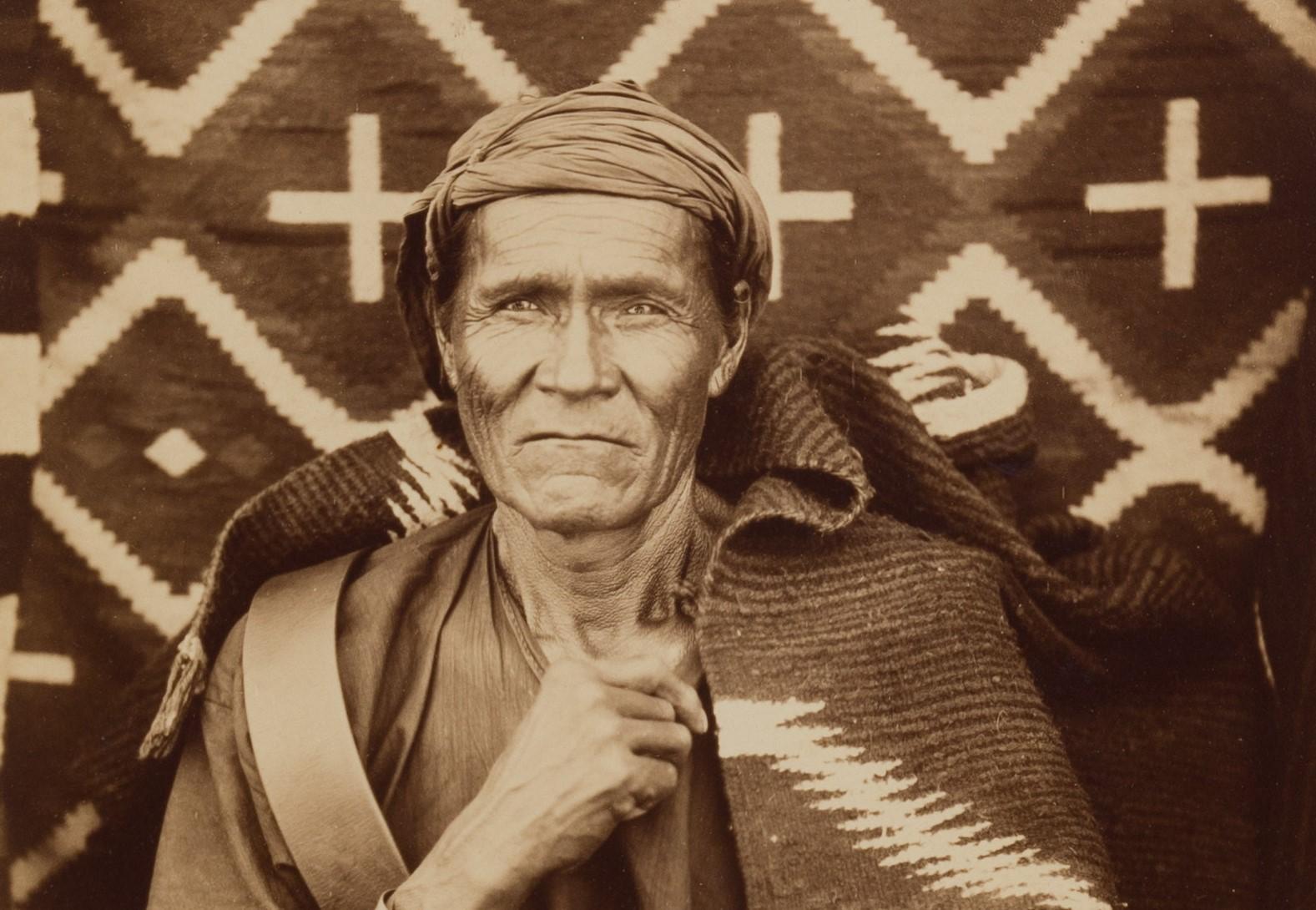 Older person described as Navajo is seated in front of a textile backdrop with geometric designs